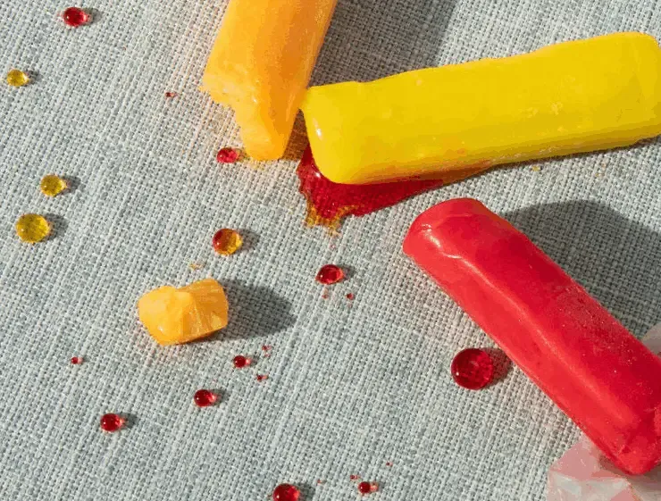 Three melted crayons in orange, yellow, and red leaving bright droplets on an off-white textured stain-resistant fabric.