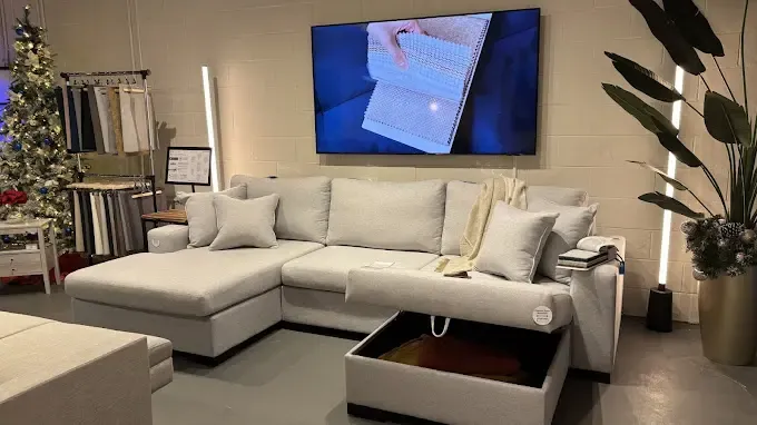 Interior of the SIXTREND Toronto showroom with a grey sectional sofa, storage ottoman pulled out to reveal interior, fabric sample rack, and a wall-mounted product video display.