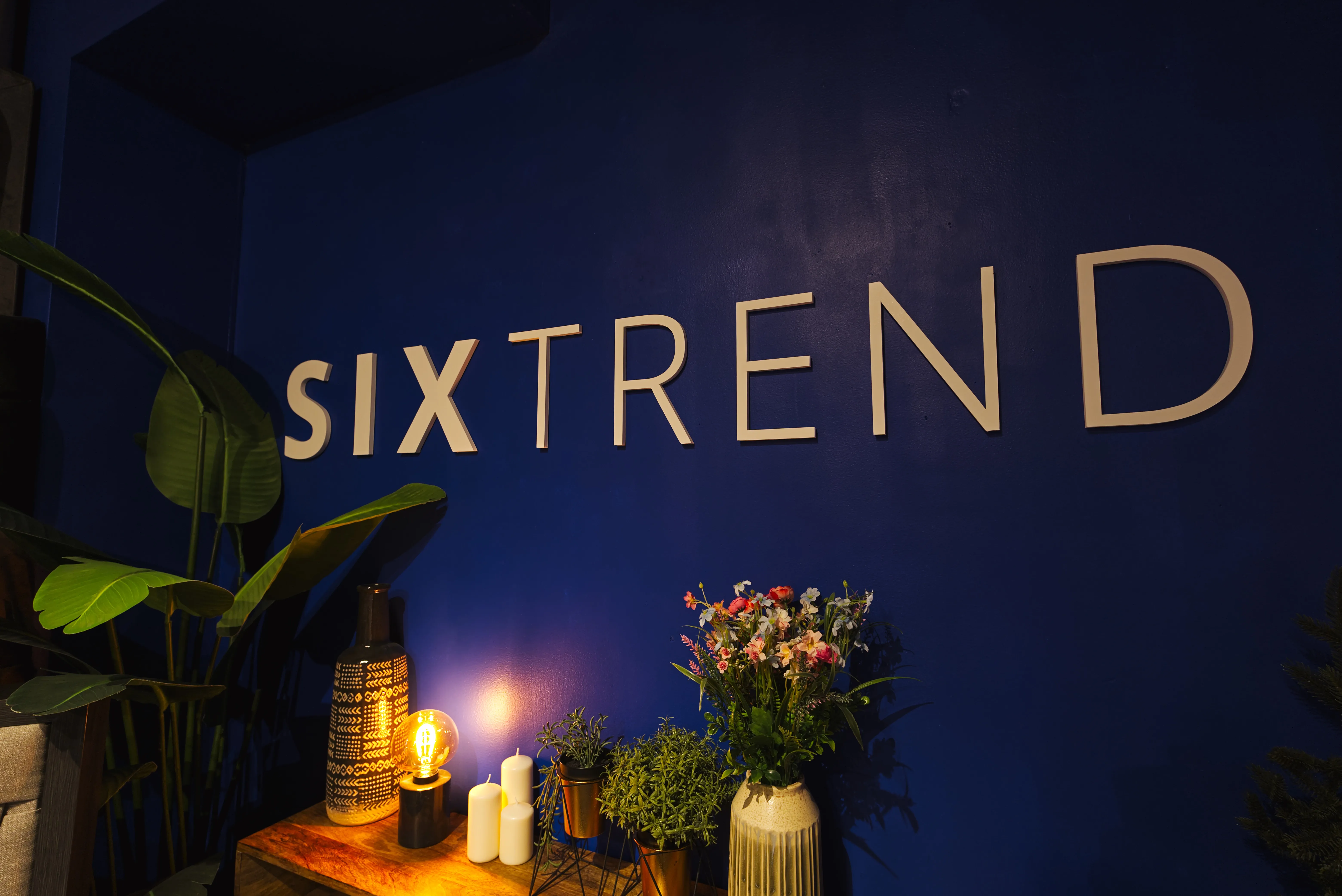 Metallic SIXTREND wordmark on a deep navy showroom wall with warm lamp, candles, and floral arrangements