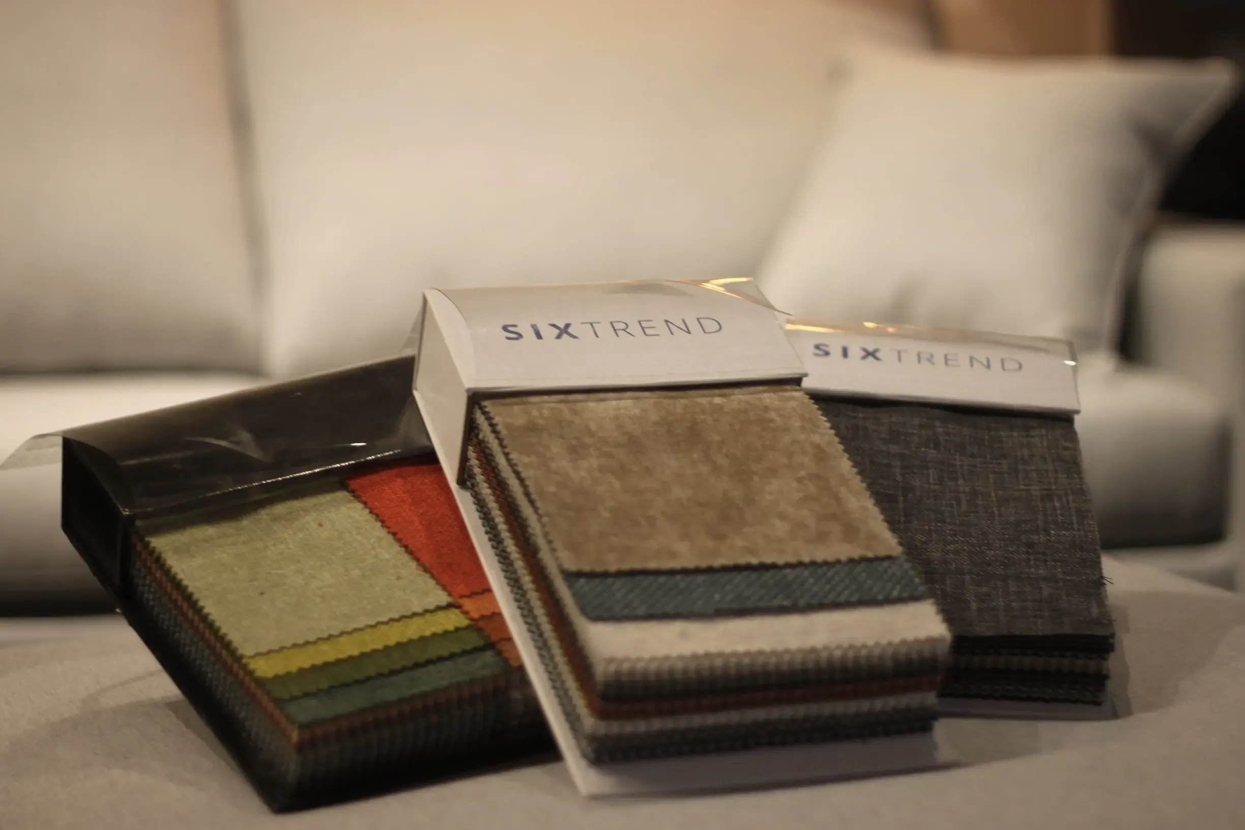 Three SIXTREND-branded fabric sample packs fanned open on a sofa, displaying layered swatches in greens, oranges, browns and grey textures.
