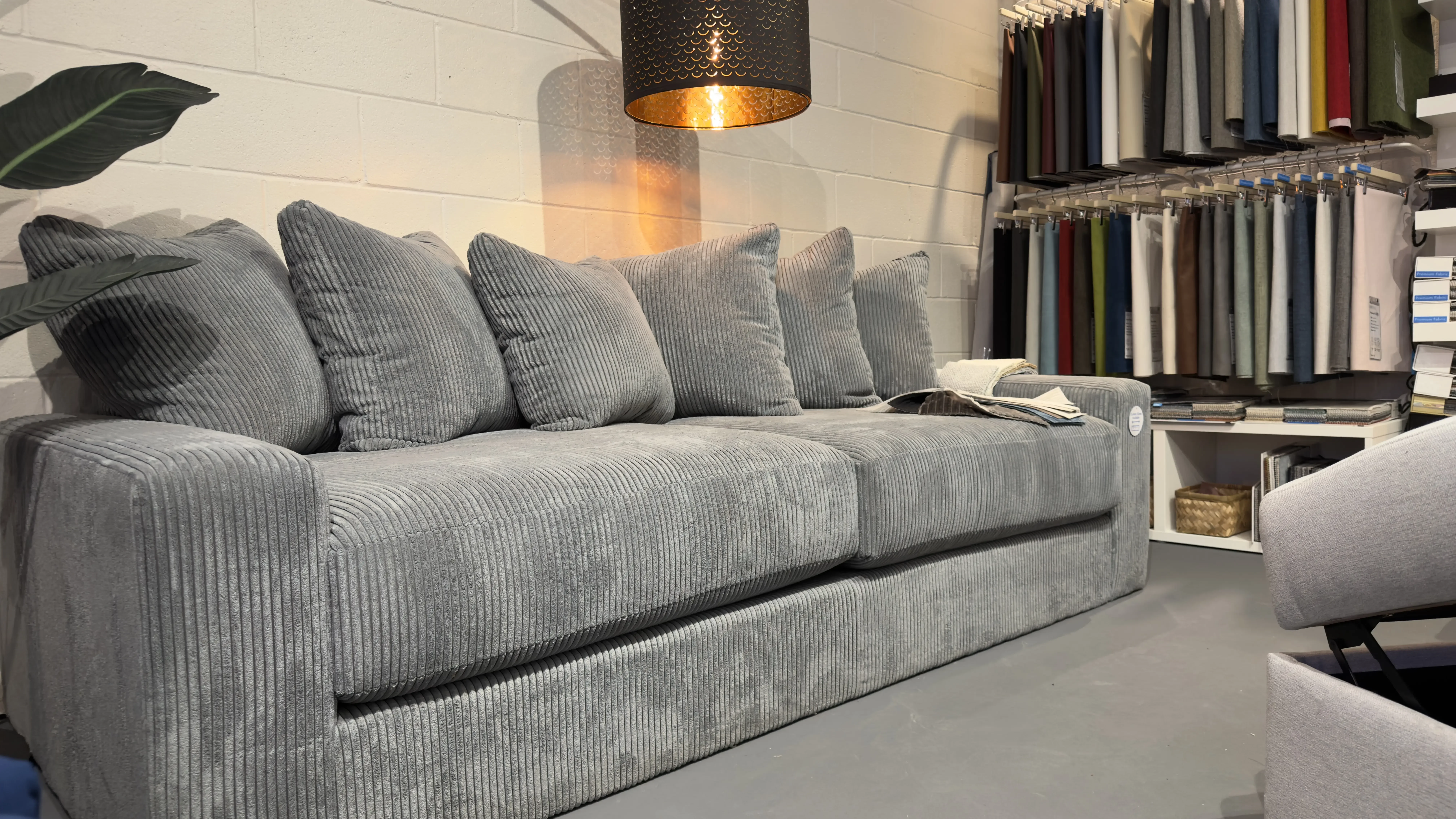 Grey corduroy sofa with deep cushions and throw pillows in a SIXTREND showroom corner, beside a wall display of upholstery fabric swatches.