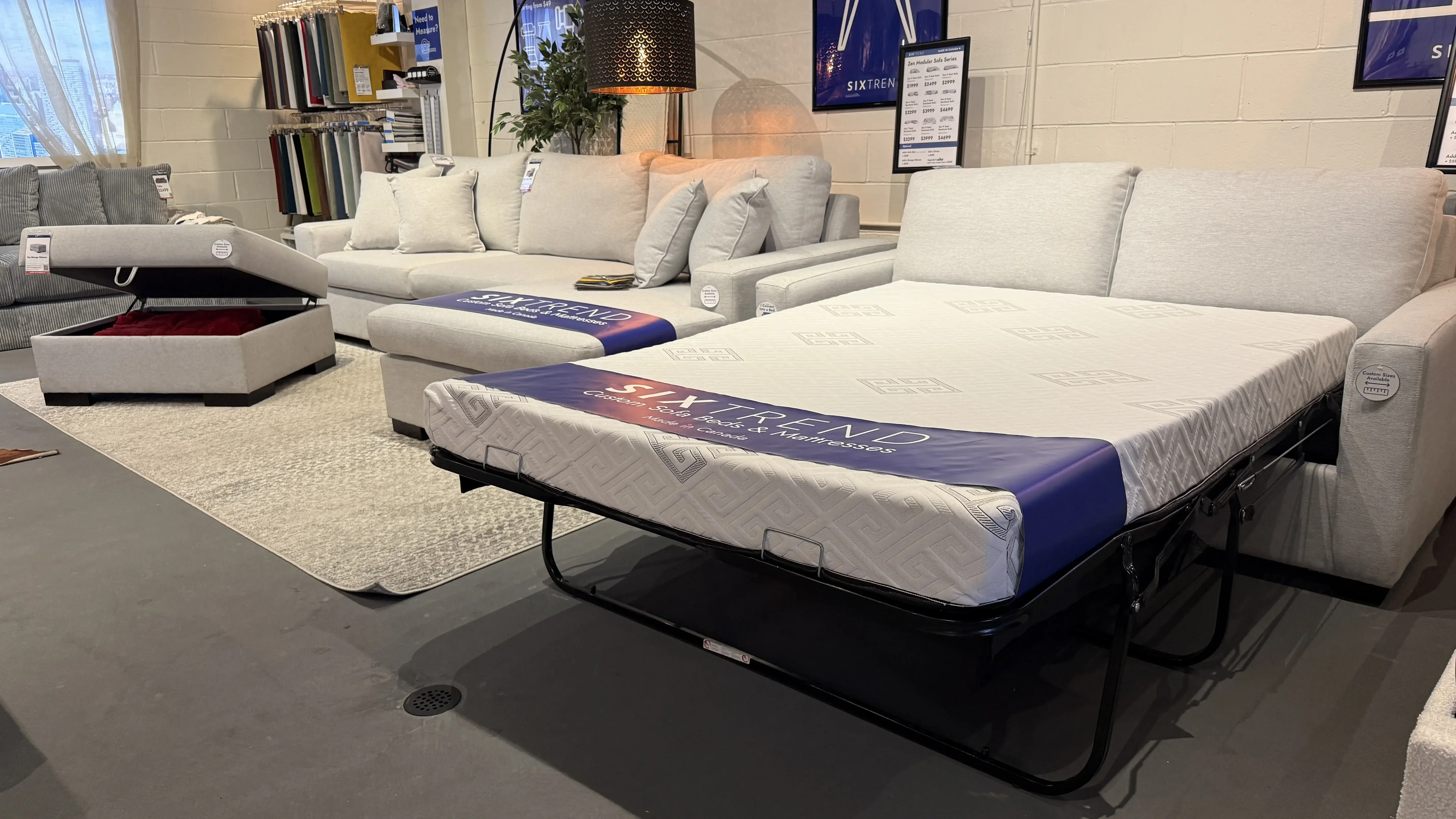 Showroom display of a light grey upholstered sofa bed with the mattress fully extended on its metal frame next to a matching sofa.