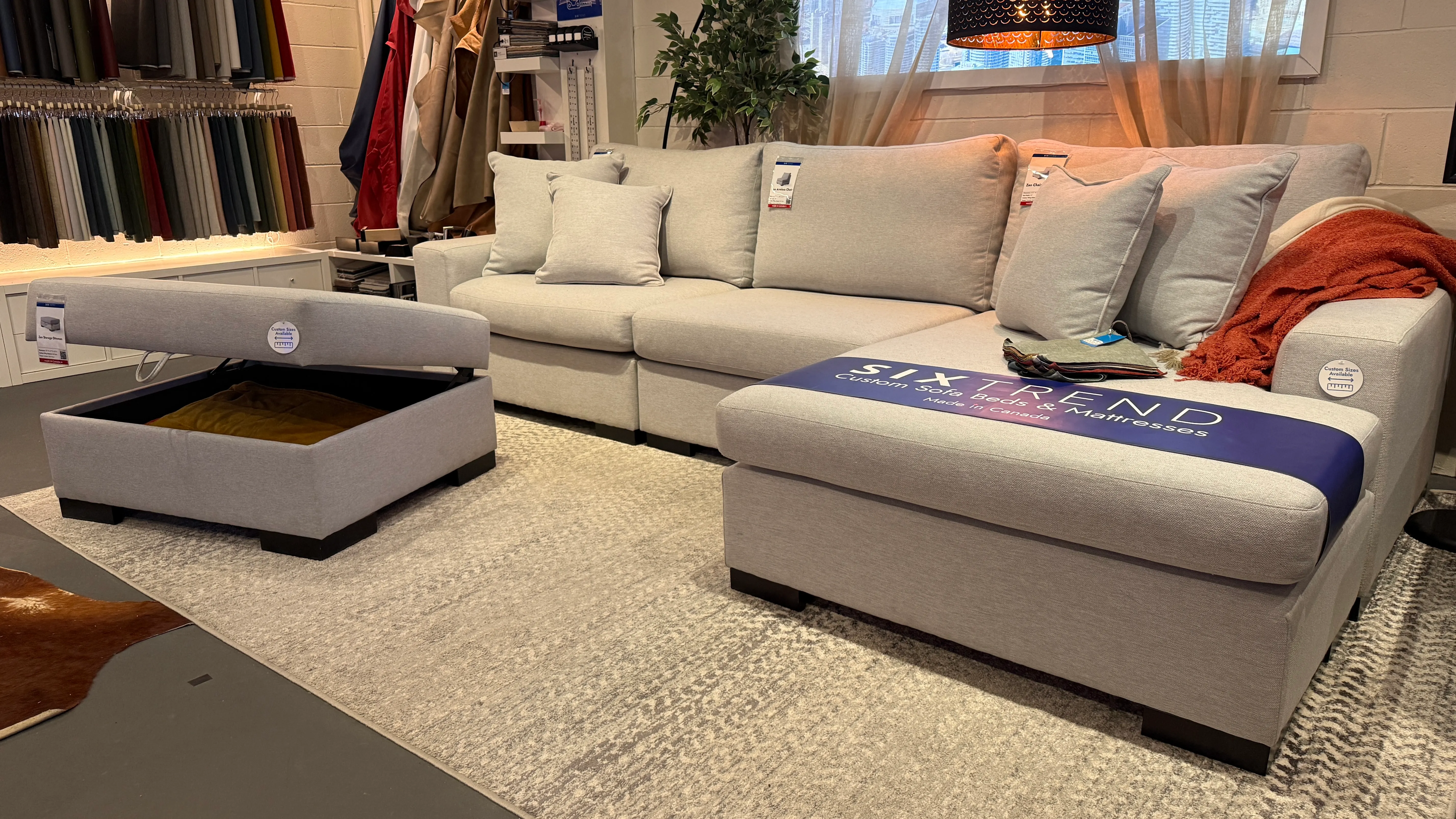 Light-grey custom sectional and storage ottoman on display in SIXTREND's Toronto showroom, with fabric swatches mounted on the wall behind